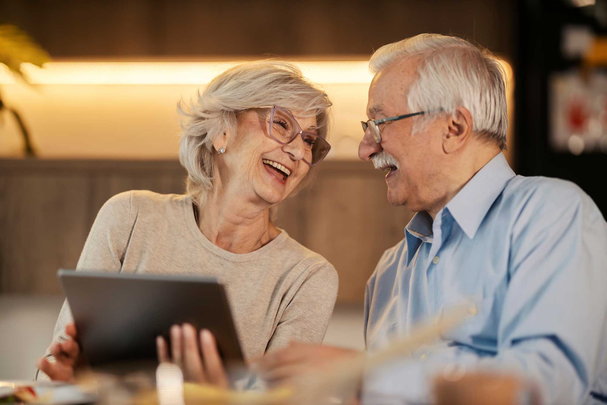 cheerful-senior-couple-laughing-at-dining-table-and-holding-tablet